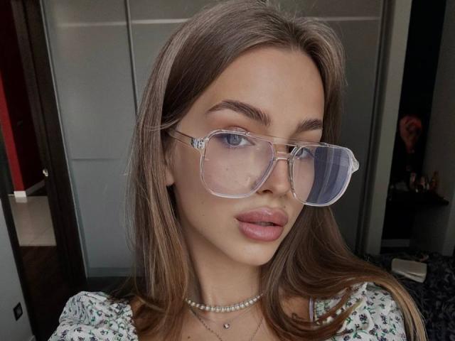 StellaHoneyX has brown hair, blue eyes, and wears clear glasses with a pearl necklace. She's pouting with glossy pink lips. She's got small tits and a cute face.