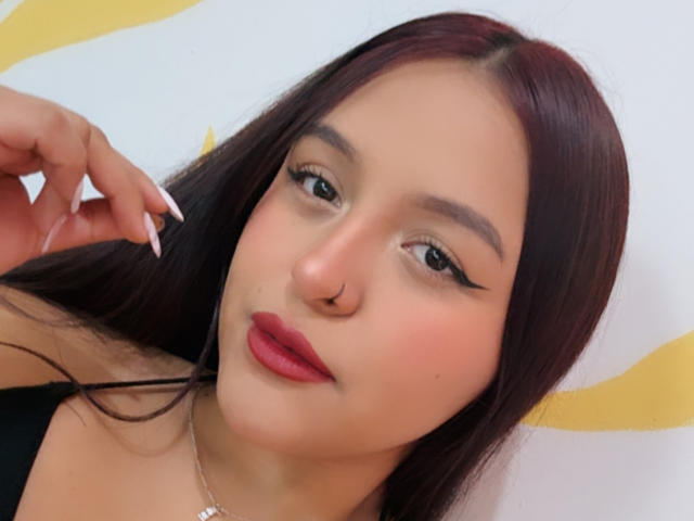MarianaVargasX heeft donkerbruin haar, rode lippenstift, zwarte eyeliner, een neusring en een schattig gezicht met lichte huid en lange witte nagels. Ze draagt een zwarte top..