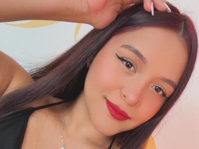 MarianaVargasX has red lipstick, black eyeliner, and long dark hair, touching her head with one hand, wearing a black top, and a silver necklace.