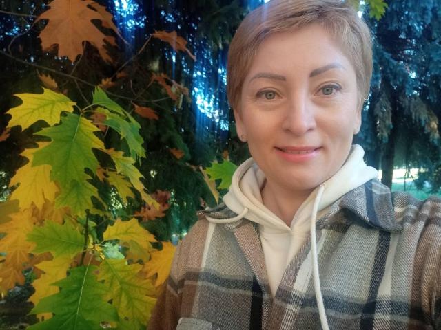 KrasaFina has short brown hair, light skin, green eyes, and a small smile. She's wearing a plaid sweater over a white hoodie, with autumn leaves in the background.