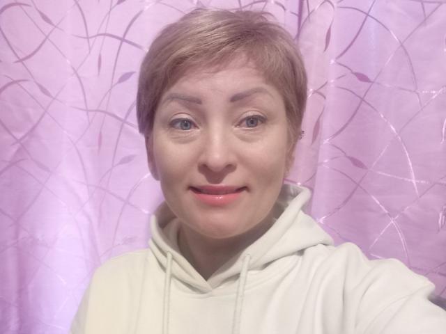 KrasaFina with short blonde hair, blue eyes, light pink lipstick, wearing a white hoodie, smiling slightly, in front of a purple curtain with abstract patterns.