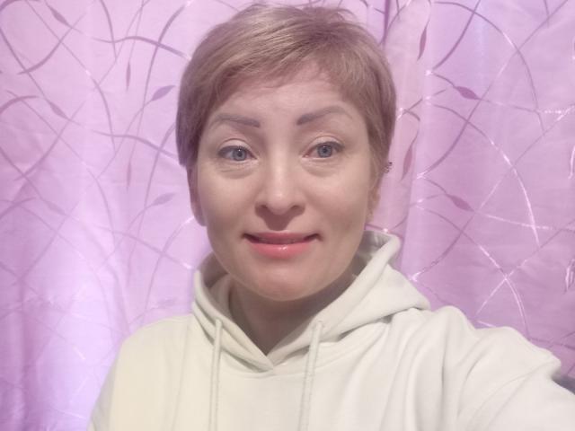 KrasaFina has short, light brown hair, blue eyes, and pink lips. She wears a white hoodie and smiles slightly. Her face is centered in the image.