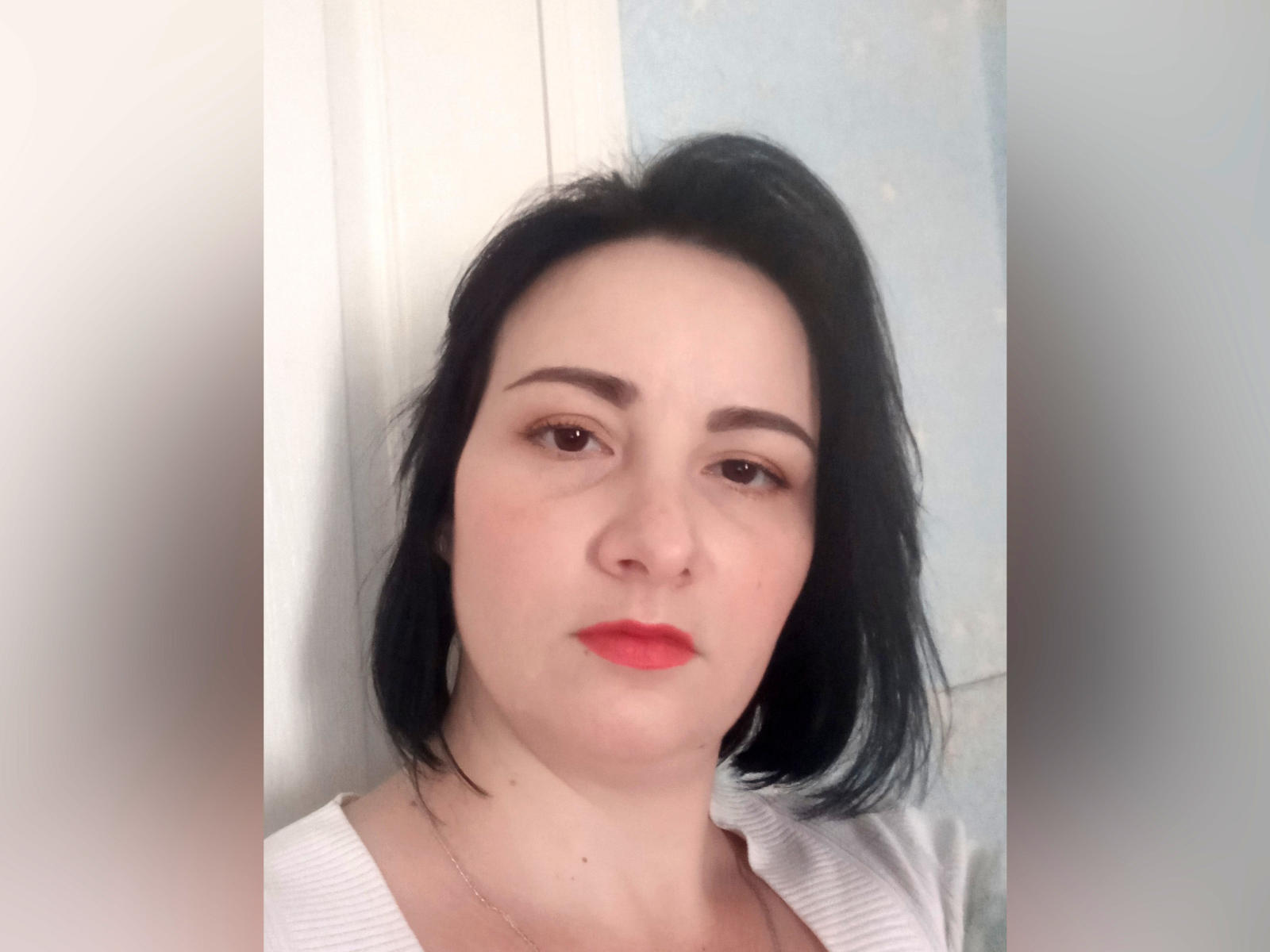 GessiNonset has pale skin, black shoulder-length hair, brown eyes, and red lipstick. She's wearing a white top, looking serious with a slight pout, and has a thin necklace.