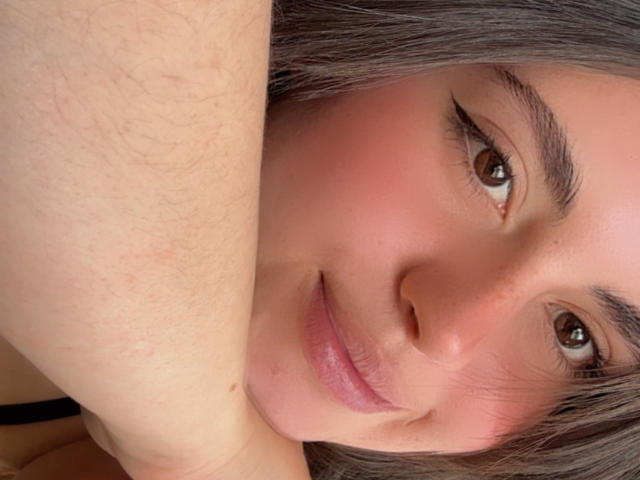 VicluX's face, showing pink blushed cheeks, brown eyes, and gray hair with a slight smile. She has a small mole near her nose and her arm is visible.