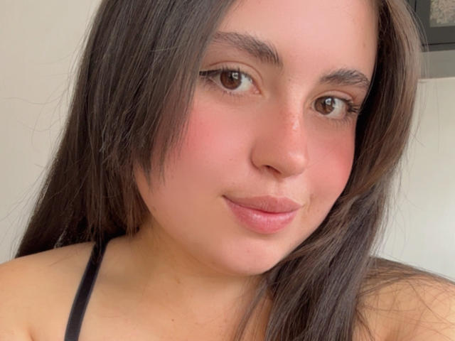 VicluX tem a pele de cor marrom clara, olhos castanhos, cabelo longo castanho e lábios rosados, vestindo uma alça preta, olhando para a câmera com um leve sorriso..