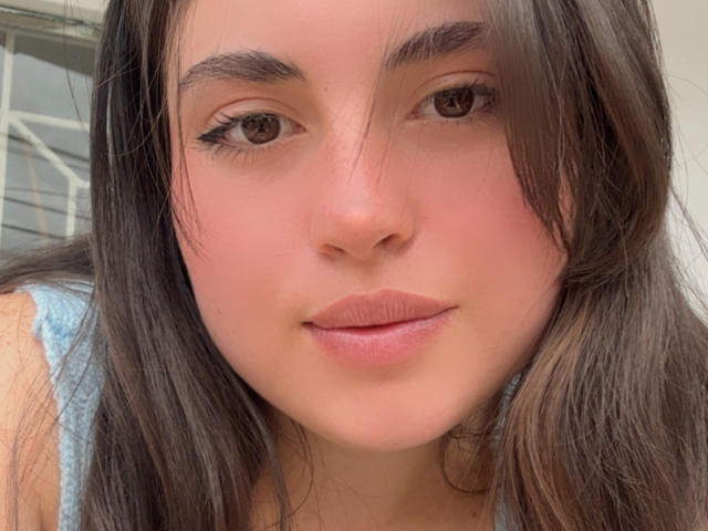 VicluX has brown eyes, long brown hair, and pink lips, wearing a light blue top. Her eyebrows are thick and she has subtle eyeliner. She's looking directly at the camera.