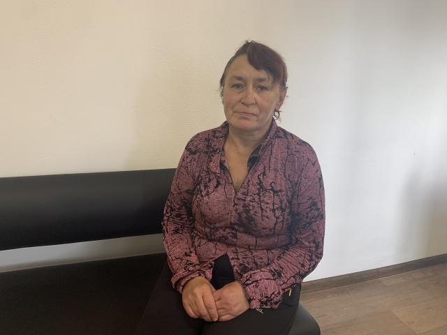 MeganSord, middle-aged woman with brown hair in a side ponytail, wearing a pink, snake-patterned blouse, sitting at a black table, hands on her lap, looking slightly annoyed.