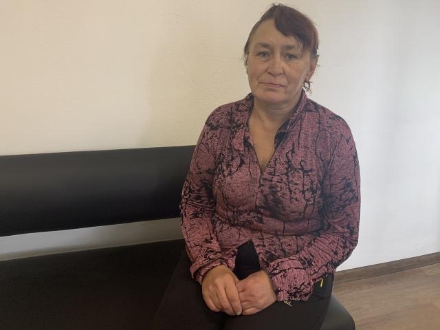 MeganSord is an older woman with red hair, wearing a pink and black patterned blouse, sitting on a black bench, hands resting on her lap, looking slightly sad.