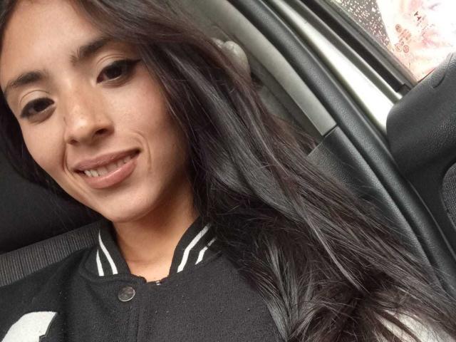AlanaMoore with long black hair, wearing a black shirt, smiling in a car, showing her small nose and slightly parted lips. Her tits are not visible.