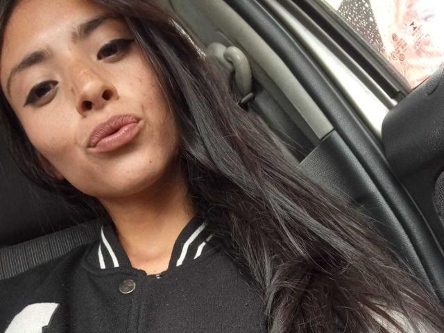 AlanaMoore has long black hair, pouty brown lips, and is wearing a black polo shirt with white stripes. She's in a car, looking at the camera.