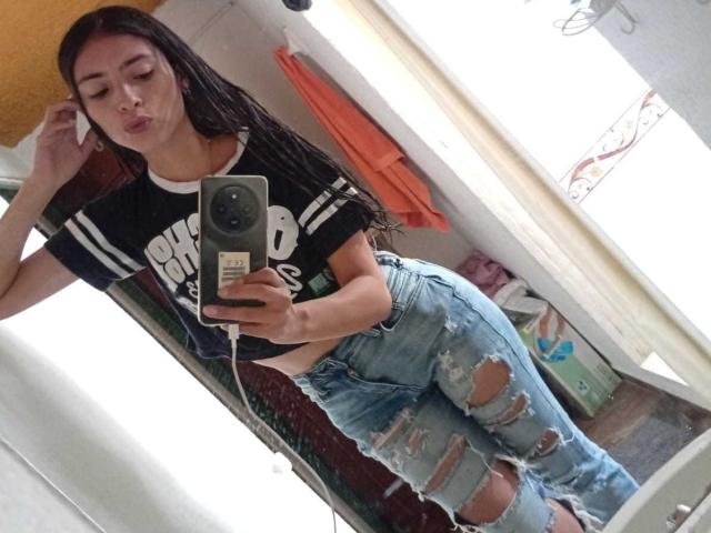 AlanaMoore with long brown hair, pouty lips, black Adidas shirt, and ripped blue jeans, taking a mirror selfie with a green camera in a bathroom.