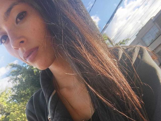 AlanaMoore has long brown hair, a serious expression, and is wearing a black zip-up hoodie with a slight visible cleavage. Sunlight highlights her facial features.