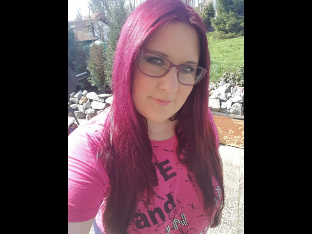JosySweet: capelli rosa, occhiali e una maglietta rosa, che mostra il seno in un selfie, sorridendo leggermente, in piedi all'aperto in una giornata di sole..