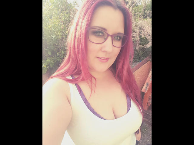 JosySweet-hot with pink hair, glasses, and a white tank top showing her big boobs, standing outside with a slight smile. Her purple bra is visible under the tank top.