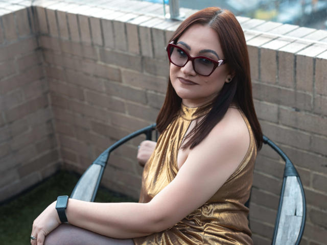 LadyNylonOh, with red glasses, brown dress, and watch, sits on a chair, showing her curvy ass and smiling at the camera. She has fair skin and straight brown hair.
