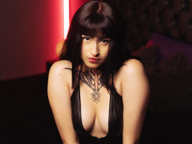 NikiEvans with dark hair and bangs, wearing a black, deep-cleavage dress, showing her tits, tattoos on chest, and a nose ring, under red lighting.