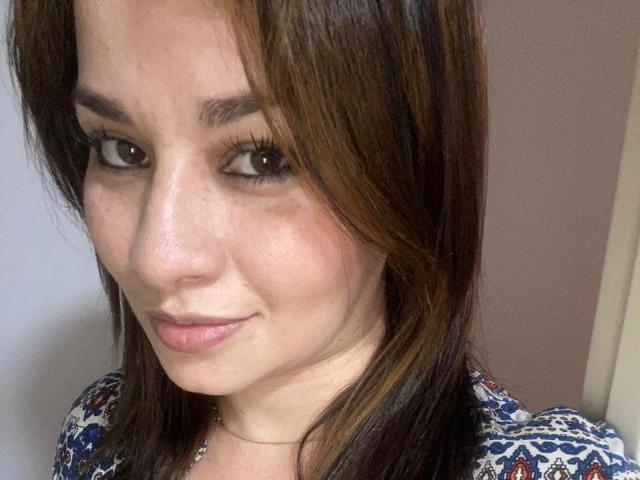 NaturalHanny has brown eyes, light skin, and straight brown hair with subtle highlights. She's wearing a patterned top, smiling gently, and has long eyelashes. Her nose is slightly shiny.