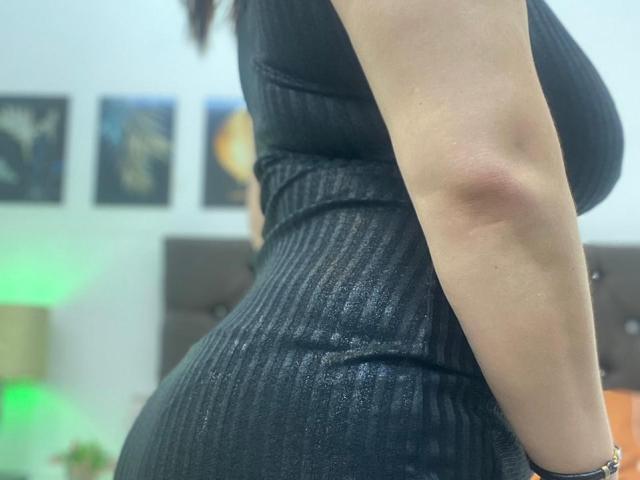 NaturalHanny's black ribbed dress hugs her curvy ass and big tits. Her right arm is visible, showing a slight tan and a silver bracelet.
