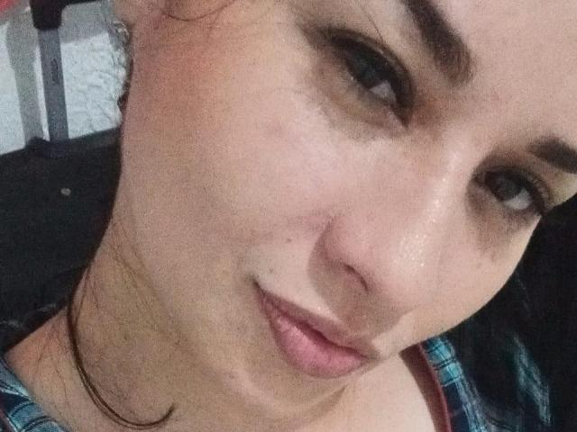 NaturalHanny's face with a small scar under her right eye, slightly parted pink lips, and brown eyebrows. She's wearing a blue plaid shirt, and has a subtle smile.