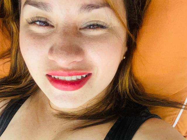 NaturalHanny's face, winking with red lipstick, black eyeliner, and subtle freckles, lying on an orange pillow, wearing a black top, with her brown hair spread out.