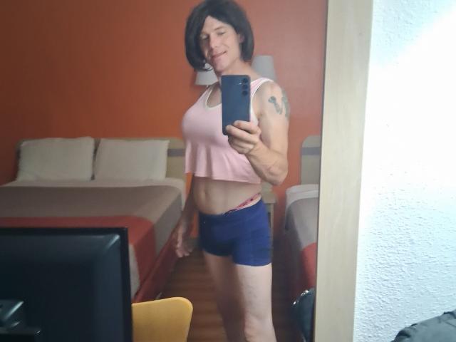 Afrodito69, avec des cheveux noirs courts, un débardeur rose et un short bleu, montrant un tatouage sur son bras droit, souriant dans un selfie devant un miroir, laissant apparaître une ceinture rose..