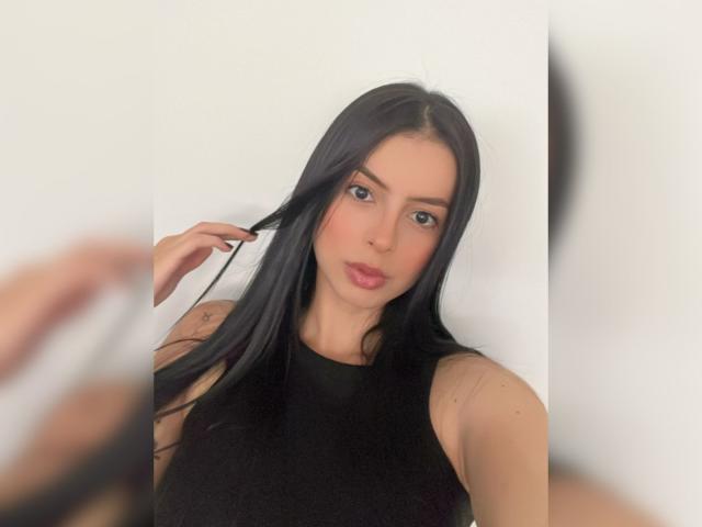 IsabellaJames has long black hair, brown eyes, pink lips, and is wearing a black sleeveless top. A hand is touching her hair. Her tits are not visible.
