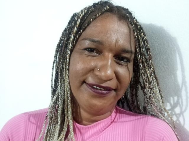 TheSecretOfRoomies heeft donkerbruine huid, gevlochten haar met blonde en bruine highlights, een neuspiercing en draagt een roze geribbelde top met paarse lippenstift..