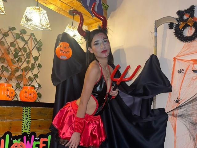 TheSecretOfRoomies in devil costume with red horns, black cape, red trident, red shiny top, pink skirt, white face paint, and black hair, standing indoors.