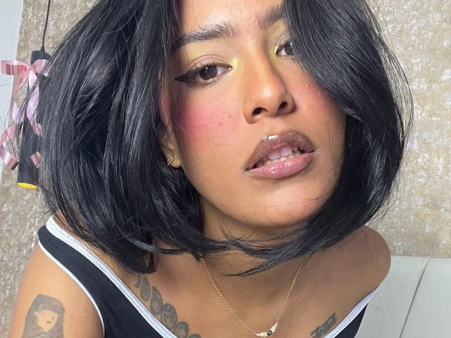 Babilett has dark, shiny black hair, pink blush on her cheeks, glossy pink lips, yellow eyeshadow, and tattoos on her chest. She wears a black tank top with white trim.