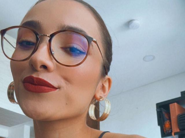 AdharaCooperr with glasses, red lipstick, blue eyeshadow, brown hair in a bun, and big gold hoop earrings, smiling slightly. Her tits are not visible.