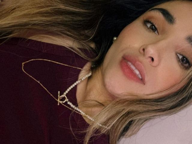 AdharaCooperr mit langem, blondem Haar, die ein dunkelrotes Oberteil und eine goldene Halskette mit einem "P"-Anhänger trägt, liegt da und lächelt mit rosafarbenen Lippen. Sie trägt ein Perlenohrgehänge..