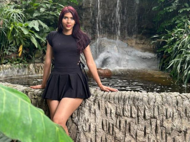 DanikaSerov with red hair, black short-sleeve top, and black skirt, leaning on textured stone, water behind, green plants in front, and waterfall in background.