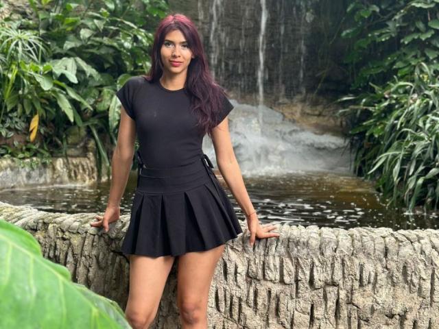 DanikaSerov with red hair, black tight shirt, and black pleated skirt, standing with hands on stone wall, water behind her, green leaves in front. Her tits are small.