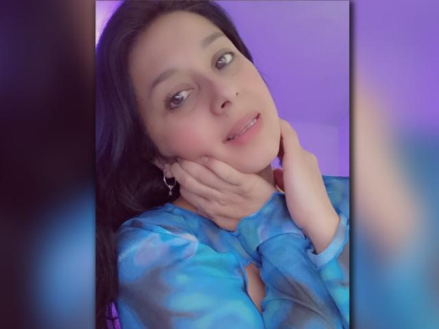 Cami69s with blue tie-dye shirt, hands under chin, smiling, dark hair, purple background, small nose ring, heart earrings, soft pink lips, slightly flushed cheeks, relaxed expression.