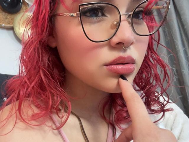 SharonFenix with red, wavy hair, wearing glasses, a nose ring, and pink lipstick, touching her bottom lip with black-nail-polished finger. She's looking at the camera.
