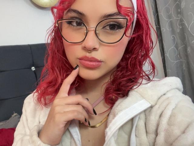 SharonFenix with red curly hair, glasses, nose ring, and pink lipstick, wearing a white hoodie, touching her chin with a finger, gold necklace, black nail polish.