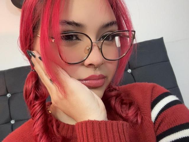 SharonFenix with red hair, glasses, nose piercings, and a red sweater, touching her face with black-nailed fingers, looking serious. Boobs are not visible.