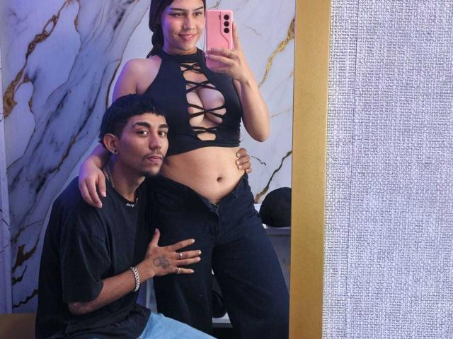 AnyelinAndMark with tits in a black lace-up top, black pants, and pink phone, stands while Mark kneels, hand on her belly, in a mirror selfie. Mark's black shirt, tattoo.