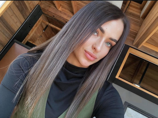 MandiBrooks with long, straight brown hair, green eyes, and pink lips, wearing a black turtleneck and green vest, smiling at the camera. Wood ceiling in the background.