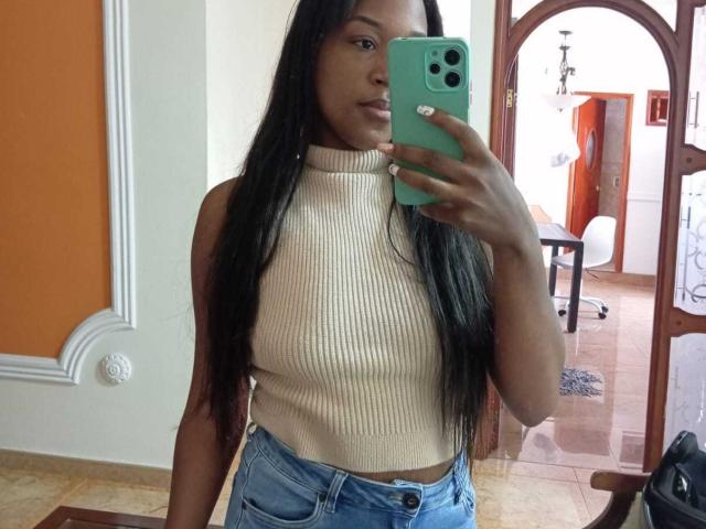 TaniaDoux with long black hair, wearing a beige ribbed sleeveless top and blue jeans, holding a green phone, showing a flat ass and small tits, taking a mirror selfie.