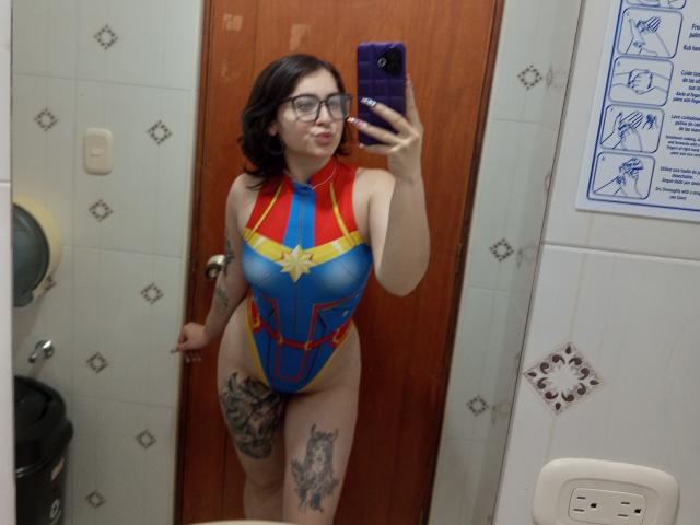 MissTat with glasses, black hair, and tattoos, wearing a blue, red, and yellow superhero one-piece, showing her tits and pussy in a mirror selfie. She's holding a purple phone.