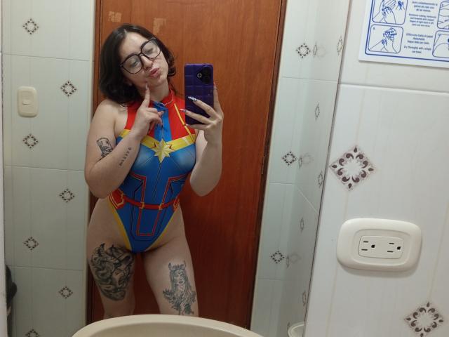 MissTat with glasses, dark hair, and tattoos, wearing a blue and yellow superhero-style one-piece, showing her tits and ass, taking a mirror selfie in a bathroom.