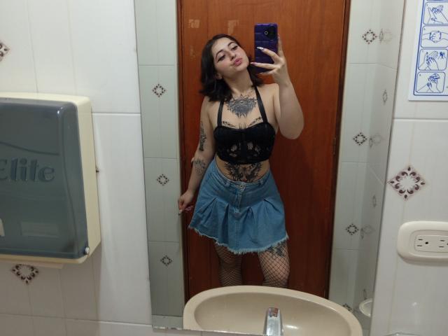 Mirror selfie of MissTat with black lace bra, denim skirt, fishnet stockings, tattoos, and dark hair, holding a blue phone, showing her tits and ass.