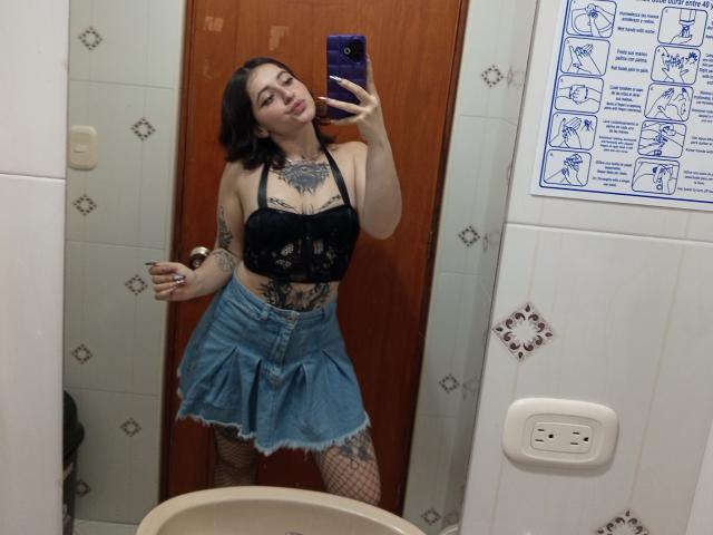MissTat with tattoos, black lace bra, blue denim skirt, fishnet stockings, holding phone, taking mirror selfie, wood door background, short black hair, red lipstick, smiling.