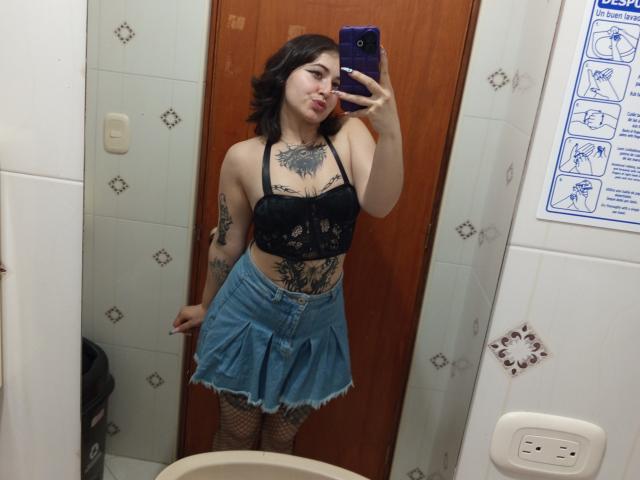 MissTat with short black hair, wearing a black lace top, denim skirt, fishnet stockings, and tattoos on her chest and abdomen, taking a mirror selfie with a purple phone.