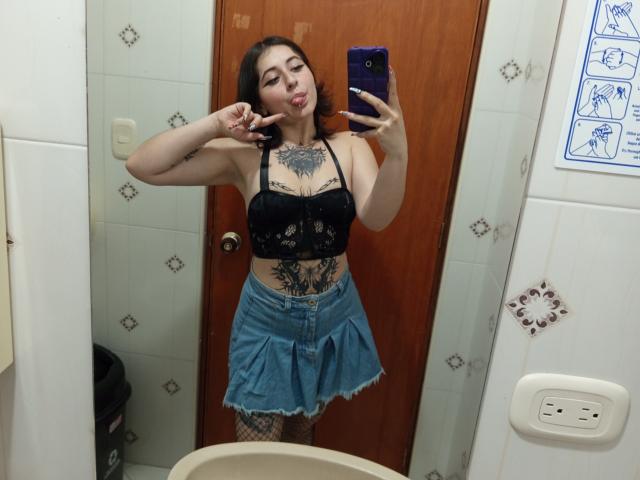 MissTat with black lace bra, blue denim skirt, and tattoos, sticking out tongue, showing off her tits and pussy tattoos, holding phone in mirror selfie.