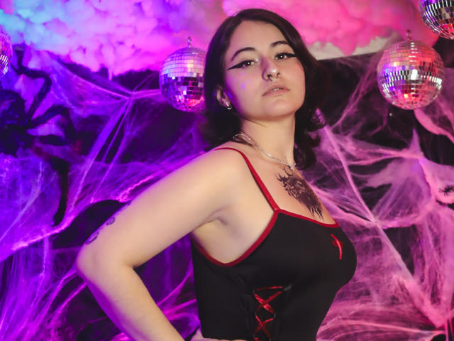 MissTat with dark hair, nose ring, and necklace, wearing a black dress with red trim, showing her tits, standing in front of mirrored balls and purple-pink lights.