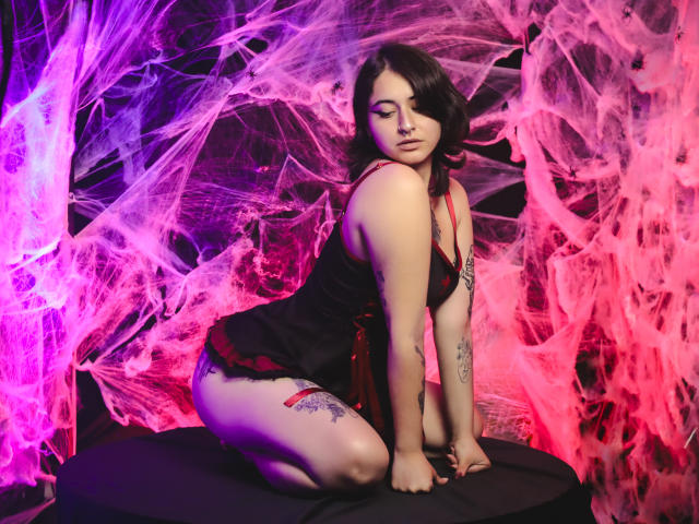 MissTat with pale skin, black hair, and tattoos, kneeling in a black and red lingerie set, surrounded by pink and purple spider webs, hands on a black surface.