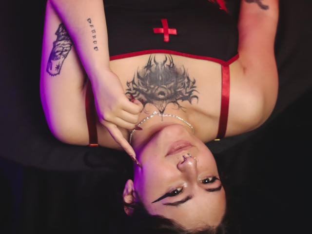 MissTat with dark tattoos, red cross dress, and nose ring, lying upside down, touching her chin. She has a wolf eye chest tattoo and a tattooed arm.