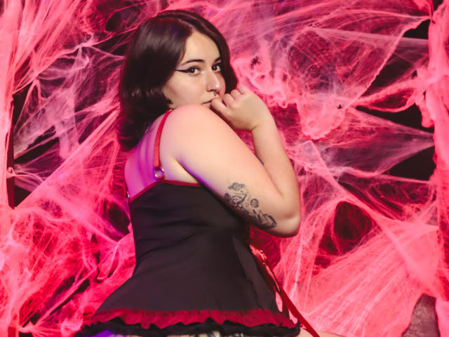 MissTat with dark hair, looking back, biting finger, black top with red straps, tattoo on right arm, pink and red net-like background, showing her ass.